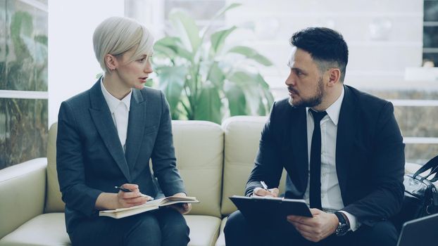 Blonde businesswoman talking to male colleague with clipboard at hotel lobby during meeting