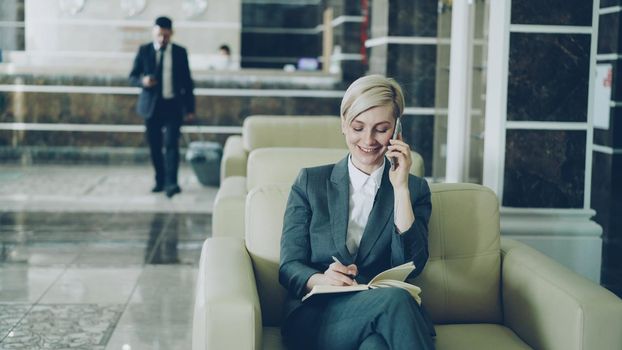 Attractive businesswoman sitting in armchair with notepad and laptop computer while businessman with luggage walking through hotel lobby from reception desk