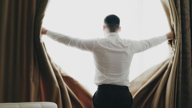 Businessman open curtains in hotel room at the morning and looking into window