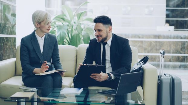 Confident businessman with clipboard talking to female colleague with notepad in hotel lobby prepraing for business meeting they arrived to. Luggage is near couch