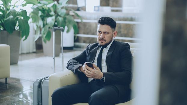 Bearded concentrated businessman using smartphone sitting on armchair inside luxury hotel after arrivel to business meeting