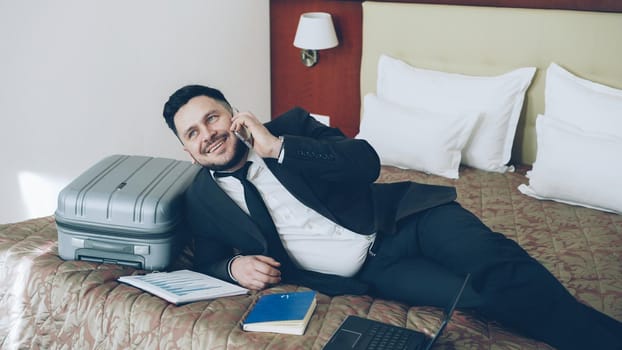 Cheerful bearded businessman talking mobile phone while lying on bed in hotel room. Travel, business and people concept