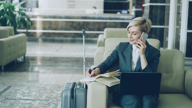 Pan shot of blonde busy businesswoman sitting in armchair in hotel lobby talking mobile phone, writing in notepad and using laptop computer smiling. Business, travel and people concept