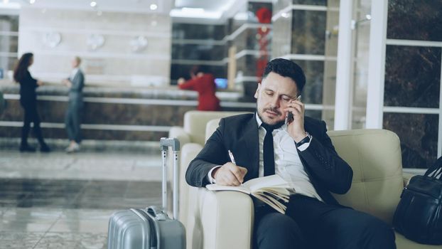 Pan shot of confident businessman talking mobile phone and writing notes in notepad while sitting on armchair in luxury hotel with luggage near him. Travel, business and people concept