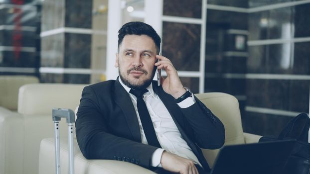 Tilt up of confident attractive businessman talking mobile phone and using laptop computer while sitting on armchair in luxury hotel with luggage suitcase near him. Travel, business and people concept