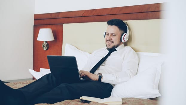 Happy cheerful businessman in headphones working at laptop computer and listening music smiling while lying in bed at hotel room. Travel, business and people concept