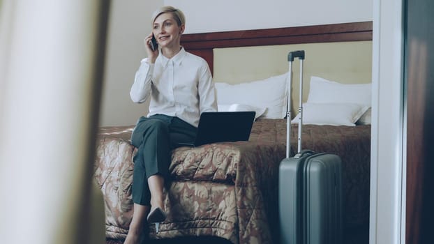 PAN of smiling blonde businesswoman in white shirt using on laptop and talking at mobile phone while sitting on bed in hotel room. Business, travel and people concept