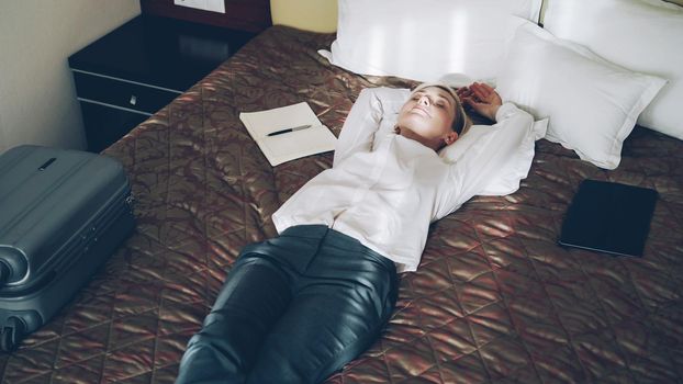 Smiling blonde businesswoman lying on bed relaxed in hotel room. Travel, business and people concept
