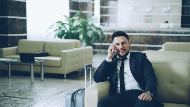 Pan shot of confident businessman talking mobile phone while sitting on armchair in luxury hotel with luggage near him. Travel, business and people concept