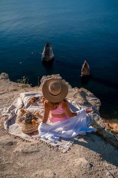 photo of a beautiful woman with long blond hair in a pink shirt and denim shorts and a hat having a picnic on a hill overlooking the sea.
