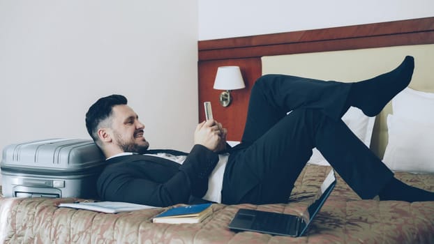 Young bearded businessman lying on bed and suitcase using smartphone while resting after arrival at hotel room. Travel, business and people concept