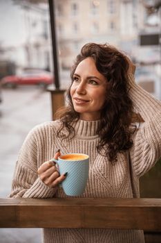 A middle-aged woman in a beige sweater with a blue mug in her hands is in a street cafe on the veranda.