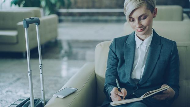 Attractive blonde businesswoman sitting in armchair in hotel lobby and writing in notepad smiling and looking aside. Business, travel and people concept