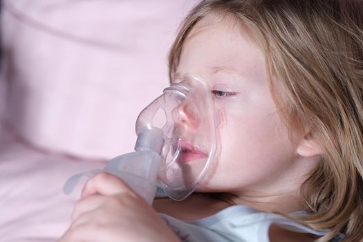 Sick girl in oxygen mask lies on hospital bed. Closeup portrait of child breathing in oxygen mask and lying in bed in pediatric ward