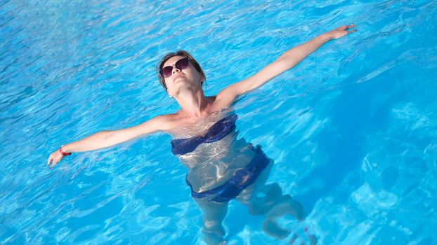 Happy beautiful woman in sunglasses relaxing in pool water. Relaxed woman in swimsuit floating in water