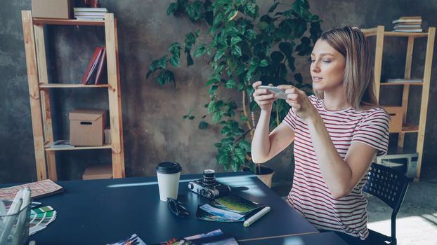 Young woman is arranging pictures, take-out coffee, sun glasses, professional camera and marker on table and shooting flat lay with smartphone, then checking photos by touching screen.