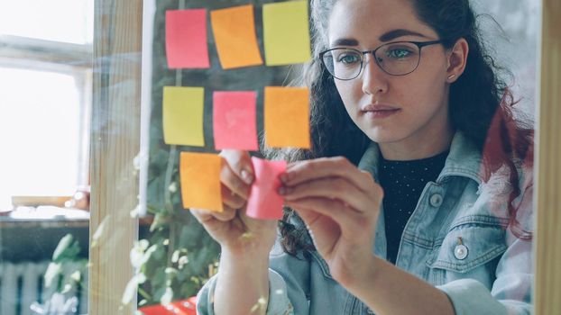 Close-up of young woman wearing glasses sticking colored memos on glass board in modern loft style office. She is looking at bright notes and smiling.