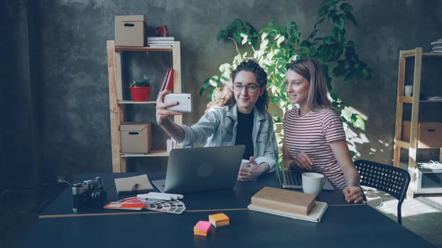 Creative young businesswomen are making selfie together in modern office while sitting at desk. They are posing with funny faces, talking and smiling