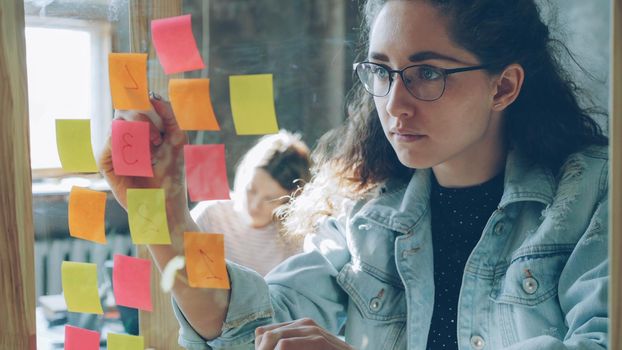 Close-up of young creative entrepreneur numbering coloured stickers on glassboard while working together with female colleague in modern office indoors