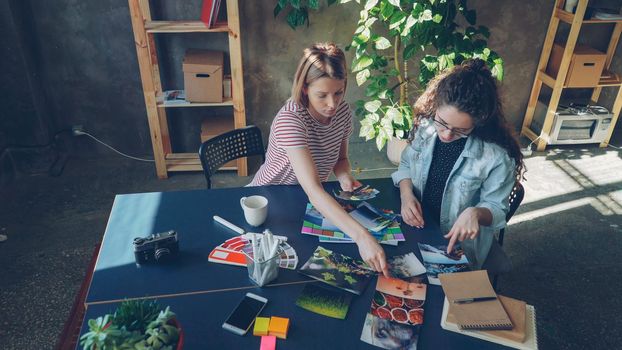 High angle view of young female designers are placing photos on broad table to create perfect flat lay working at studio. They sitting at desk together and discussing different photo