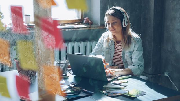 Creative young businesswoman is listening to music in headphones dancing while working at desk with laptop in modern office indoors. Glass with colored stickers in foreground.