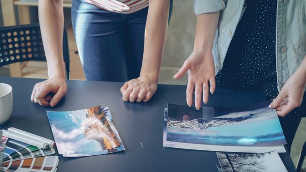 Tilt-up shot of women's hands placing photos on modern table in modern office . Female designers are grouping pictures by color and image and discussing illustrations.