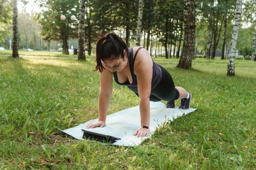 A charming brunette woman plus-size body positive practices sports in nature. Woman does yoga in the park on a sports mat. On a yoga mat in the dog pose.