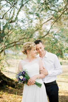 Groom hugs bride from behind with a bouquet near the olive tree. High quality photo