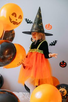 Children's Halloween - a girl in a witch hat and a carnival costume with airy orange and black balloons at home. Ready to celebrate Halloween.