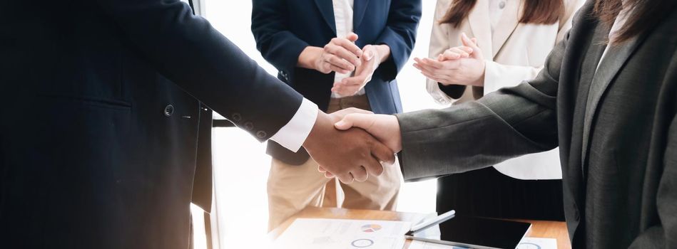 Two business men shaking hands while their colleagues applauding in the background.