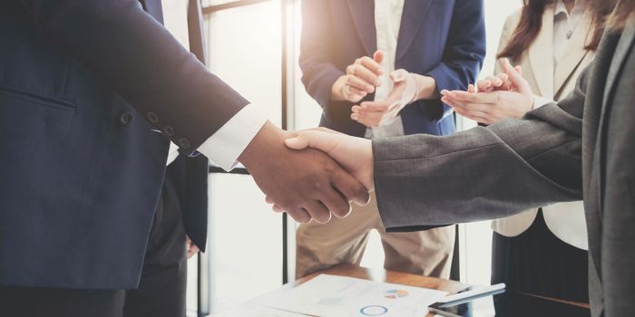 Two business men shaking hands while their colleagues applauding in the background.