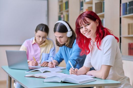 Smiling teenage female student looking at camera while sitting at desk with classmates in library. High school, college, knowledge, education, study, youth concept
