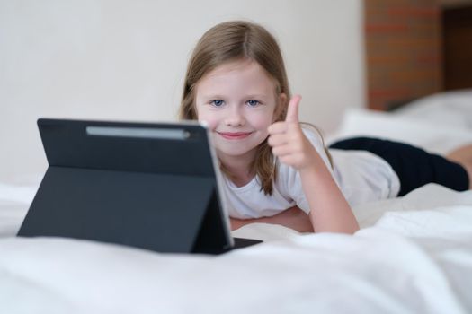 Cheerful child girl showing thumb up lying on bed with tablet. Children rest at home with modern gadgets