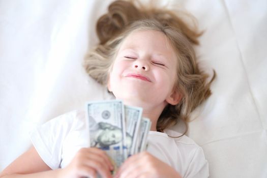 Happy girl holding cash dollars lies on bed with eyes closed. Childish dreams and lot of money