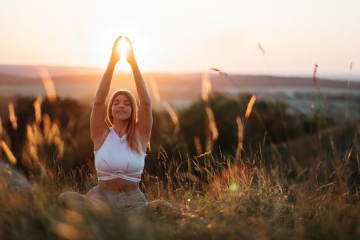 Young Woman Sitting in Meditation Yoga Pose and Catching Sun by Hands at Sunset Outdoors