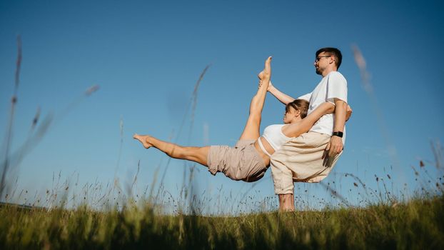 Man and Woman Dressed Alike Doing Difficult Pose While Practicing Yoga Outdoors in the Field with Blue Sky on Background