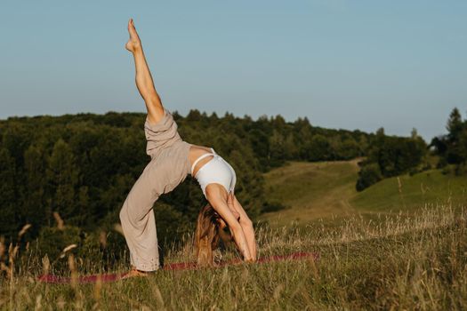 Young Woman Practicing Yoga Outdoors in Field with Blue Sky on the Background and Scenic Landscape at Sunset