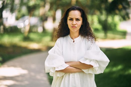 Angry emotions of a curly-haired brunette girl with crossed arms in the park. High quality photo