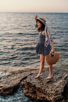 A woman in a dress, hat and with a straw bag is standing on the beach enjoying the sea. Happy summer holidays.