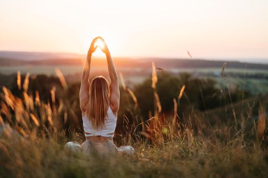 Back View on Woman Sitting in Meditation Yoga Pose and Catching Sun by Her Hands at Sunset Outdoors