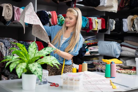 A blonde woman inspects a sheet of clothing blanks while holding them in her hand. Planning the start of the tailoring work of the new collection.