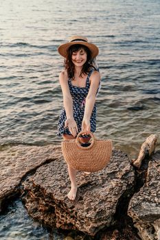 A woman in a dress, hat and with a straw bag is standing on the beach enjoying the sea. Happy summer holidays.