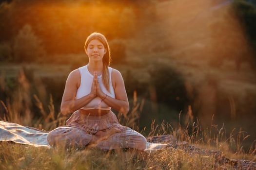 Happy Young Woman with Folded Hands and Closed Eyes Sitting on Mat and Meditating Outdoors at Sunset
