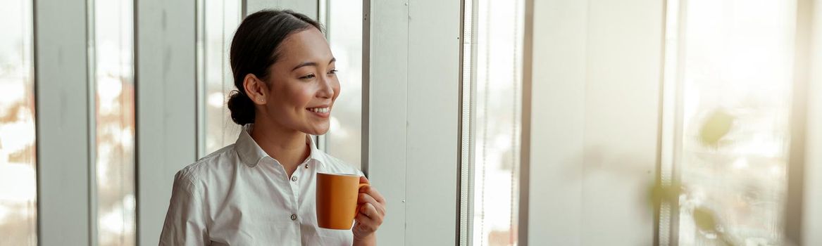 Portrait of asian business woman drinking coffee standing near windows at office. Blurred background