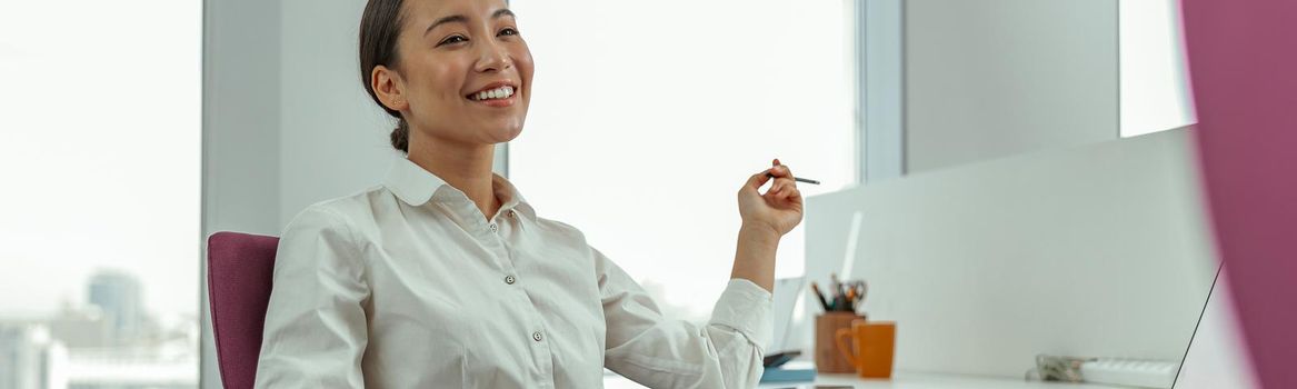 Smiling asian business woman sitting in office on her workplace and looks away. High quality photo