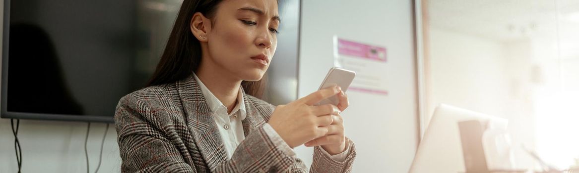 Upset asian business woman using phone while working on her workplace in office. High quality photo