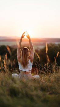 Back View on Woman Sitting in Meditation Yoga Pose and Catching Sun by Her Hands at Sunset Outdoors