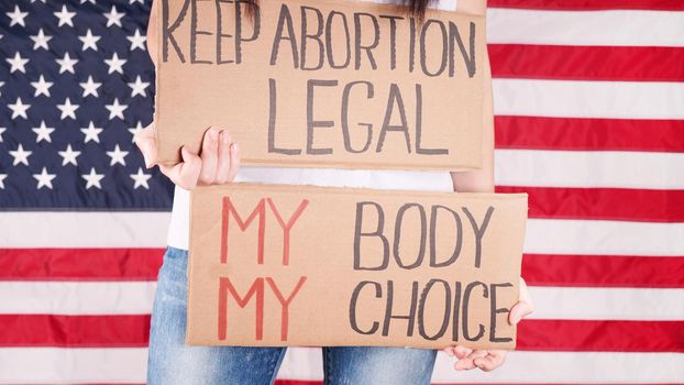 Young woman protester holds cardboard with Keep Abortion Legal and My Body My Choice signs against USA flag on background. Girl protesting against anti-abortion laws. Womens rights freedom