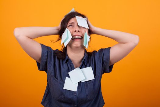 Portrait of overwhelmed business woman with work in studio over yellow background. Woman with sticky notest on her head. Blue sticky notes.