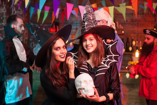 Beautiful young witch holding a human skull and smiling to the camera at halloween party. Witches looking into the camera.